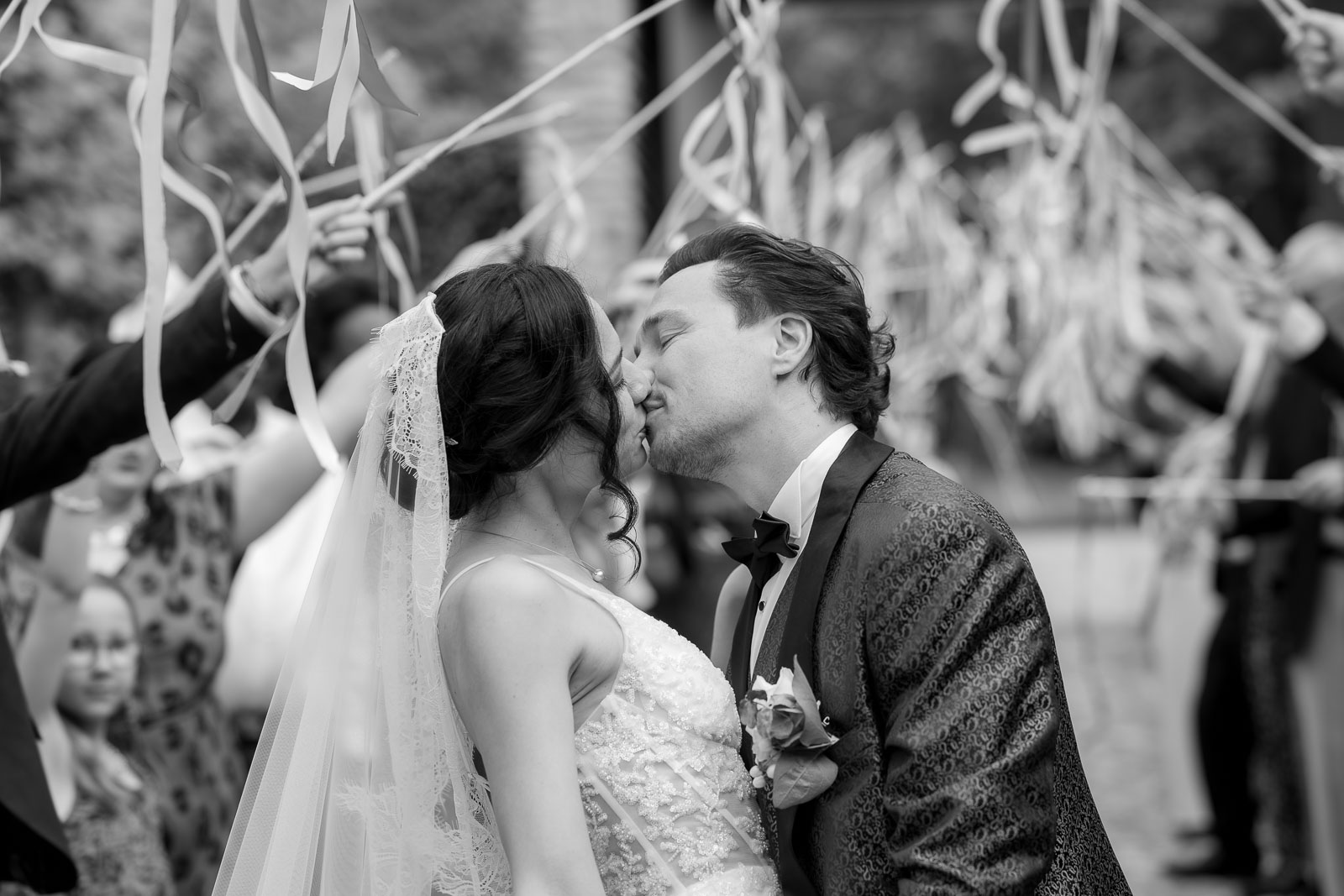Photos du mariage et Morgane et Bruno – Bruxelles-73 Baiser des mariés sous une haie de rubans à la sortie de la cérémonie, en noir et blanc